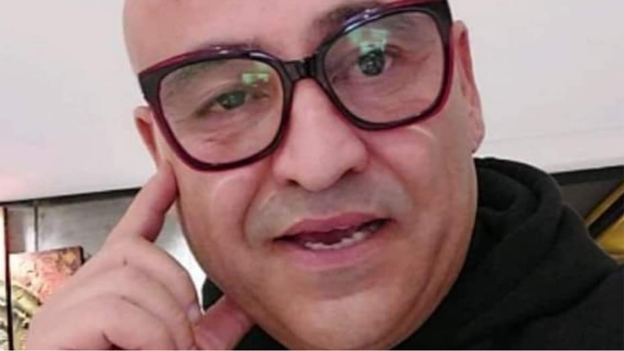 A person with large glasses and a shaved head is touching their cheek and looking at the camera. They are wearing a black top and are indoors.